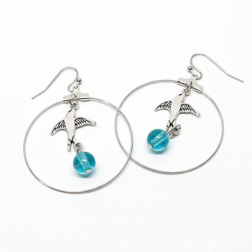 Silver hoop earrings with bird charms and blue beads on a metallic surface